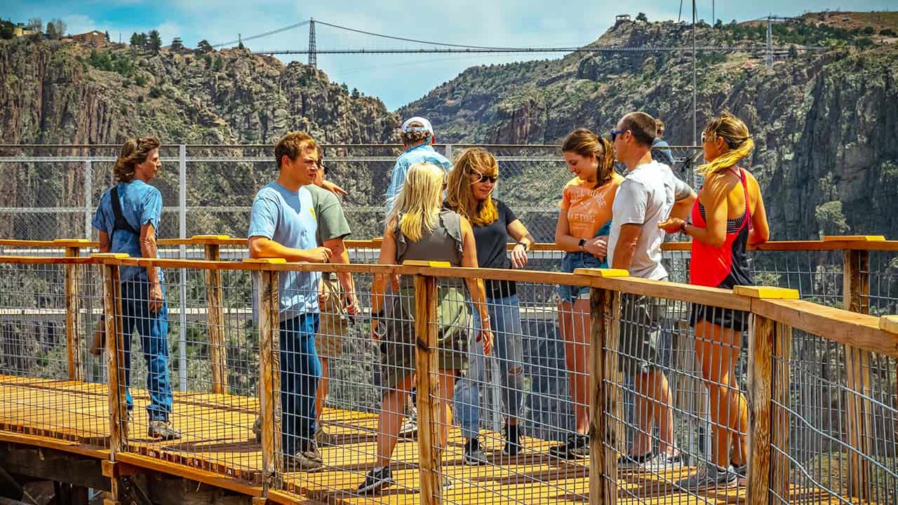 A group of visitors on a wooden overlook above the Royal Gorge, with the suspension bridge spanning the canyon behind them