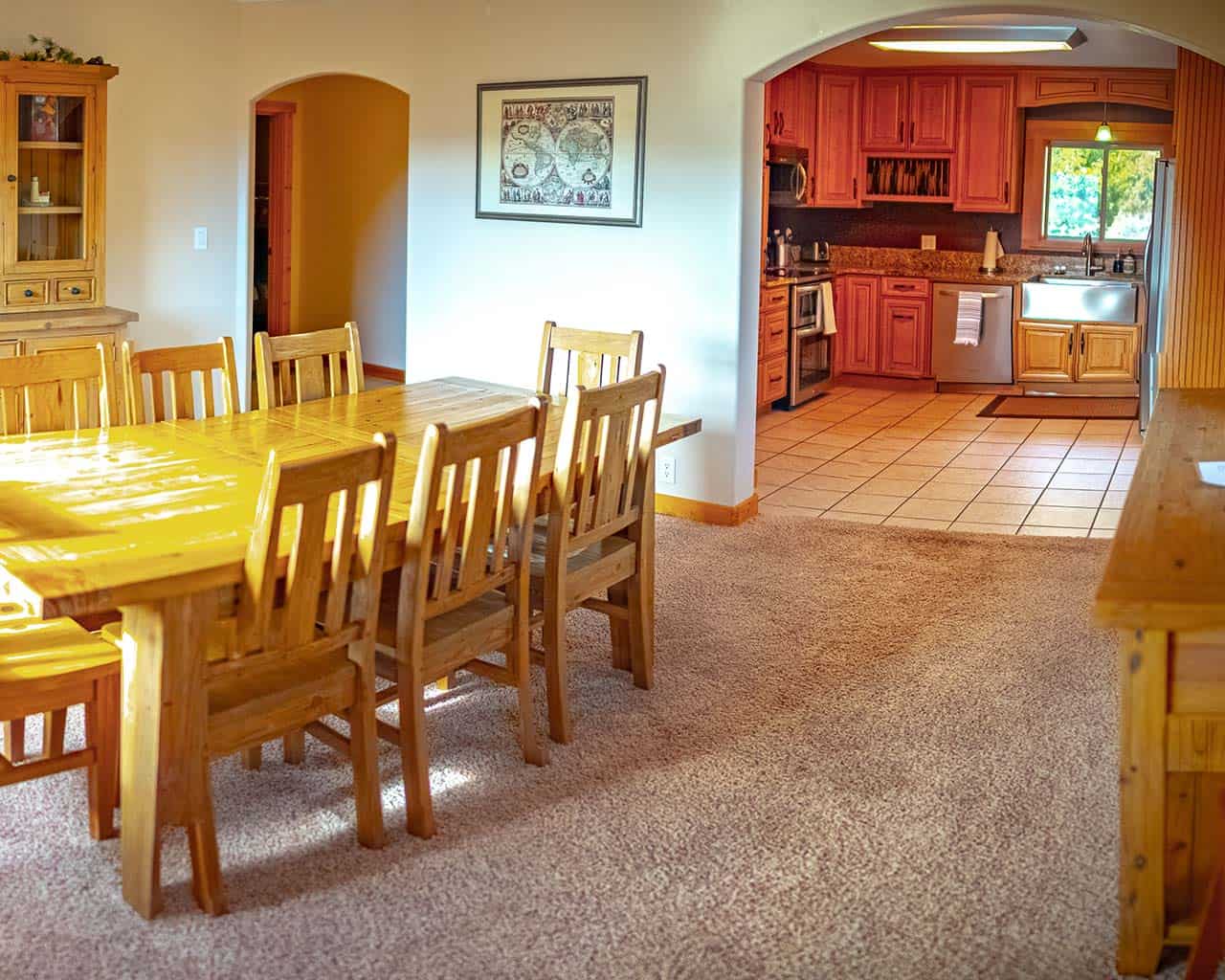 A long wooden dining table and chairs, opening through an arched doorway into a kitchen with wood cabinets